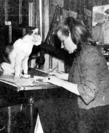 Black and white image of a woman (right) working in a studio while her cat (left) sits on her desk