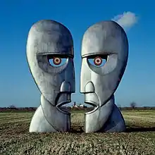 A colour photograph of two large silver-grey iron sculptures of opposing silhouetted faces. The sculptures are standing in a brown wheat field with a blue sky behind them.