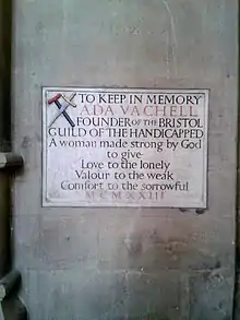 Engraved plaque. At the top left is a crutch crossed by a sword, the symbol of the guild of the handicapped she founded. The plaque reads: A woman made strong by God to give love to the lonely Valour to the weak Comfort to the sorrowful