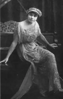 A young white woman seated on a piano bench, wearing a headband with a white lacy ruffleand a long light-colored lace gown with loosely draped short sleeves