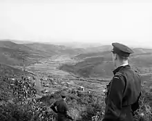 Man in dark-coloured military uniform and peaked cap looking over a valley