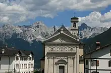 A wide angle shot of Rivamonte Agordino with a church in the foreground alongside some European style buildings. The background is slightly out of focus and is dominated by a landscape of rocky mountains with green hills and a clear sky above it.