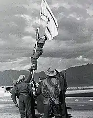 Israeli soldiers stabilize a flag pole whilst another soldier climbs it in order to raise an improvised flag; the soldier is seen about halfway up the flag pole. Other soldiers look on.