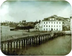 Photograph of a long wooden bridge crossing a river to a town with multi-storey white buildings and palm trees in the far background