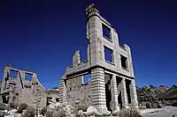 Ruins of a three-story masonry building rise into a cloudless, dark blue sky. The building is roofless, and large sections of its walls are missing. Masonry rubble lies about the building, which has openings for doors and windows but no glass or wood. Another ruin is nearby, and barren hills are visible in the distance.