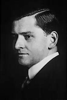 A black and white portrait of a man in a suit and tie. Half of his face is in a shadow.