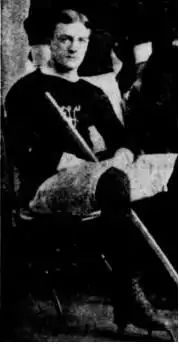 Black-and-white photo of a sitting man wearing a sweater-like hockey jersey and holding a hockey stick