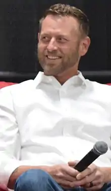 A 42-year-old man with brown hair and a brown beard smiling to the left of the camera.