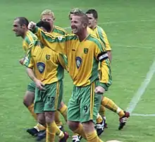 Five men standing on a grass football pitch, wearing yellow shirts, green shorts and yellow socks.