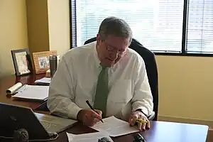 A grey-haired man with a white shirt and green tie sits at a desk in front of an open window, looking down to follow his pen as he writes on a sheet of paper