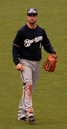 A man wearing a navy blue jersey with "Brewers" on the front in white, gray pants, navy blue cap with a white "M", and outfielder's glove on his left hand walking on a grassy field