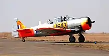 A low-winged monoplane pointing from left to right. Both of its tandem canopy places are open, showing the two aircrew, and its two-bladed propeller is still. It is bare-metal with high-visibility orange markings, and the symbols of the SAAF.