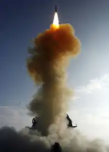 At top, a dark rocket is emitting a bright plume of flame against a blue sky. Underneath, a column of smoke is partly concealing a navy ship.