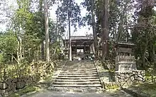 Staircase  leading to a wooden roofed gate. Trees on either side of the staircase.