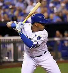 A man in a white baseball jersey and blue helmet prepares to swing at a pitch.