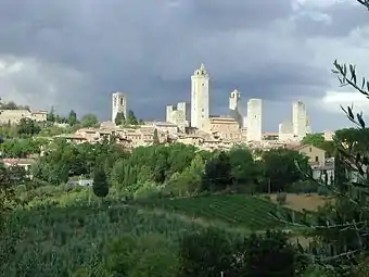 View of a small town on a hilltop surrounded by trees and vineyards. There are eight tall square towers rising from among the densely packed houses.