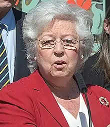 Sandy Galef, a white woman with white hair and wearing a red jacket, stands in front of a microphone.