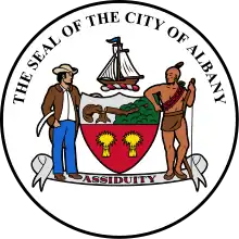 Circular seal with central images of a shield at center and sailing ship above it, with a European man to the left and a Native American to the right. The seal's edge reads "THE SEAL OF THE CITY OF ALBANY" with "ASSIDUITY" in a banner above the bottom.