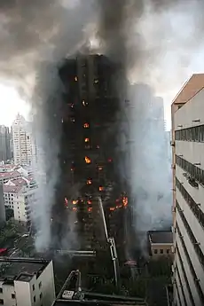 Flames can be seen inside a tall brown-black building frame. Smoke surrounds the gutted structure while a single stream of water from the ground reaches midway up the building.