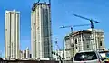 Ground level view of three towers; the one farthest to the left is complete, and has an all-white facade with balconies and light windows. The two buildings on the right are both under construction; the middle tower is nearly complete, with some concrete still exposed and a crane on the roof. The tower farthest to the right is also incomplete and has about 10 stories of framework constructed.