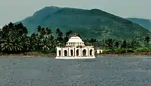 A view of a pond with a hall like structure built in the middle, surrounded by green trees and the hills in the background.