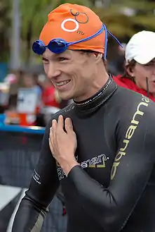 A man wears a black long-sleeved wetsuit, an orange swimming cap, and blue-shaded swimming goggles. He is smiling and holds his left hand over his chest.