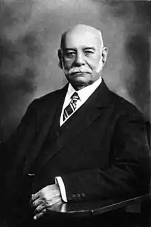 Solemn studio portrait of a distinguished bald gentleman with white sideburns wearing a 3-piece suit; left arm on armrest of a chair