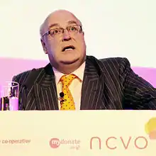 Photograph of a man in a suit standing behind a podium so only his upper chest and head are visible. His mouth is open as he is speaking.