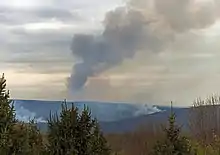 A distant ridge, seen as spring appears to be emerging, with two areas from which white smoke is rising. In the center a large plume of smoke, dark against a cloudy late afternoon sky through which some sunlight is penetrating, rises and bends to the right. In front are some evergreens.