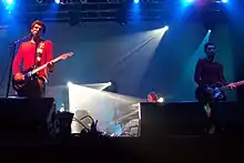 Two men playing a guitar in a concert, with another playing keyboards in the background.