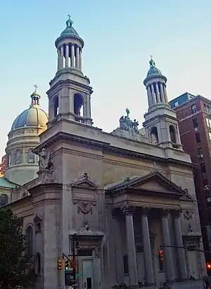 A stone building with a pediment and columns on the main entrance, two towers with green rounded tops and columns, and a dome at the rear lit by late afternoon sun from the right. There are traffic lights in front.