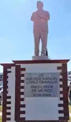 Picture of the statue of López Obrador, a stone sculpture placed on a pedestal. López stands while he embraces himself.