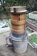 A simple hearth with a metal pan holding two wooden steaming vessels and a wooden lid used in Japan.