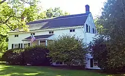 A white house with gray roof and large shrubs in front flying an American flag from the porch