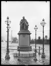 A black and white photograph of a statue atop a plinth surrounded by a low fence and several tall lightposts