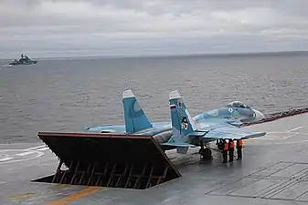 Rear port view of aqua-and-white jet aircraft lining up on aircraft carrier's deck, preparing for takeoff. The jet blast deflector is erected behind the aircraft. Three men in bright orange fluorescent tops stand underneath the jet's right wing