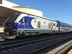 A diesel locomotive with navy blue curved shapes on the front and rear with white accents, a black cab area, and Amtrak California logos on the front and sides