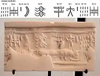 A modern impression of an Indus cylinder seal discovered in Susa, in strata dated to 2600-1700 BCE. Elongated buffalo with line of standard Indus script signs. Tell of the Susa acropolis. Louvre Museum, reference Sb 2425. Indus script numbering convention per Asko Parpola.