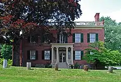 A brick building with a flat roof, dark green wooden window shutters and yellow wooden trim including a small entrance portico with Greek columns behind a large tree on its left. There is a row of five wooden stumps in front.