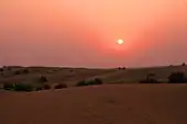 View of dunes at sunset. The setting sun hangs low and dim in a sky graded from fiery orange at top to maroon near the horizon. Terrestrial features are difficult to discern in the crepuscular illumination.