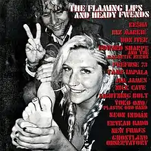 A black-and-white photo of Kesha holding her foot to the camera with a smiley face drawn on her toe, standing in front of Wayne Coyne who is giving a peace sign with his fingers and smiling. The title of the album and the collaborators are written in red to the right.