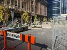 A street with dirty, damaged cars and broken concrete, behind an orange and black "Road Closed" sign.