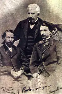 An old photograph depicting two dark-haired men seated in the foreground and a white-haired man standing behind