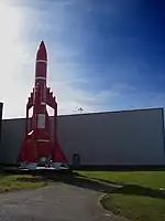 A model of a rocket-shaped spacecraft with three engine nacelles around its cylindrical main body. Photographed standing upright in front of a building.