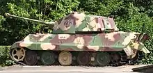 The side of a large tank, freshly painted in pale yellow, green and rust-brown camouflage, sitting in sunlight on a concrete plinth.