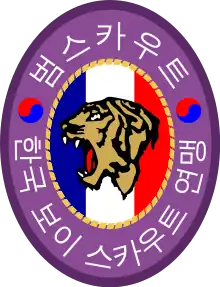 An oval badge of red with 범스카우트 (Tiger Scout) and 한국 보이 스카우트 연맹 inscribed in hangul; The center is an oval of yellow with the profile of a tiger head over a scroll inscribed with "preparation" and knot