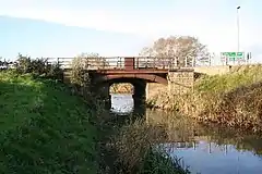 A narrow drain crossed by a small bridge. The bridge is of rusted, and untreated steel, and consists of a straight steel beam supporting the road deck, with, behind it, a steel arch. The arch is fixed in a buttress of dressed and coursed stonework, visible only on the far bank.  There is another on our side of the drain, but it is obscured by vegetation. Rusty handrails protect pedestrians from falling.  The water is flat, reflecting (where it is not in shadow), the blue sky above.