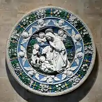 A circular terracotta plaque, sculptured in relief and glazed in intense colours of blue and green with white figures and motifs. At the centre the Virgin Mary, watched by John the Baptist, kneels in adoration of the baby Jesus. Little cherubs look on