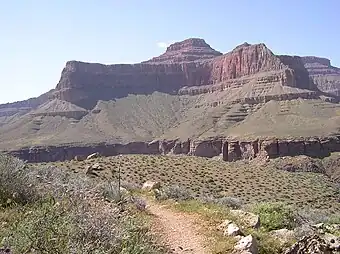 View of Tower of Set peak and sub-unit cliff section from Tonto Trail, Granite Gorge, north of Mohave Point, Grand Canyon Village, South Rim. The peak is behind and separated from a cliff unit (with small prominence), in front-(photo center, right, Tower of Set (peak) to its left). Vertical erosion in cliff of Redwall Limestone, upon horizontal Muav Limestone cliff. The Tapeats Sandstone sits in foreground on Granite Gorge,  and is seen as thinly-bedded. The slope-former above is the (dull-greenish)-Bright Angel Shale with thin, inter-bedding, as well as one resistant cliff unit. The Redwall Limestone cliff section in Grand Canyon is about 450 feet (137 m) thick.