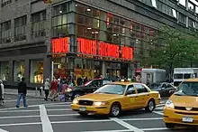 A street view of a store front prominently features a yellow taxi in front of the store. The walls contain clear windows with the phrase "Tower Records - Video" in bright orange lights.